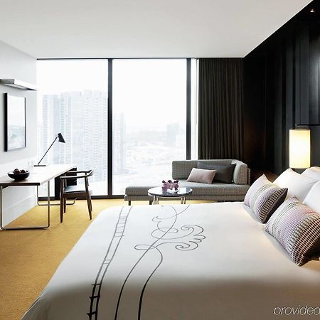 Hotel Crown Metropol Melbourne Camera foto The photo shows a modern hotel room featuring a large bed with decorative pillows, situated against a dark wall. There is a desk with a chair next to a window that offers a view of the cityscape. A small seating area includes a couch and a round tabl