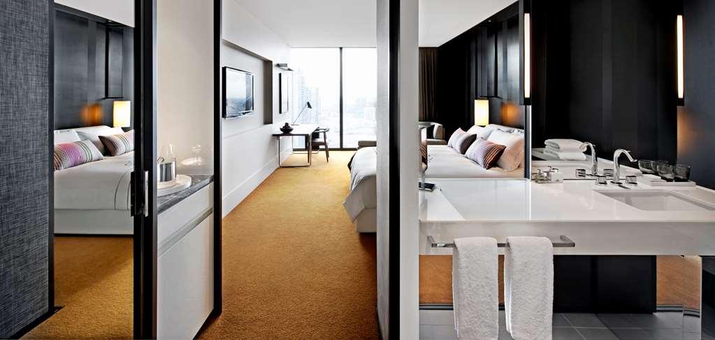 Hotel Crown Metropol Melbourne Camera foto The photo shows a modern hotel room with a sleek and stylish design. On one side, there is a sleeping area with a large bed, adorned with decorative pillows, and two lamps on bedside tables. Across from the bed, a flat-screen TV is mounted on the wal