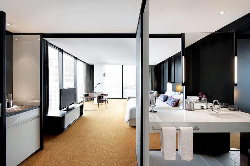 Hotel Crown Metropol Melbourne Camera foto The photo shows a modern hotel room that features an open design. On one side, there is a spacious sleeping area with a large bed, stylish bedding, and bedside tables. Adjacent to the bedroom, there is a living space with a desk, a comfortable chair,
