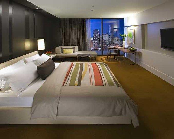 Hotel Crown Metropol Melbourne Camera foto The photo depicts a modern hotel room featuring a king-sized bed with a white and gray comforter and decorative pillows. There's a large window with a view of the city skyline at dusk, allowing natural light to fill the space. The room has dark walls