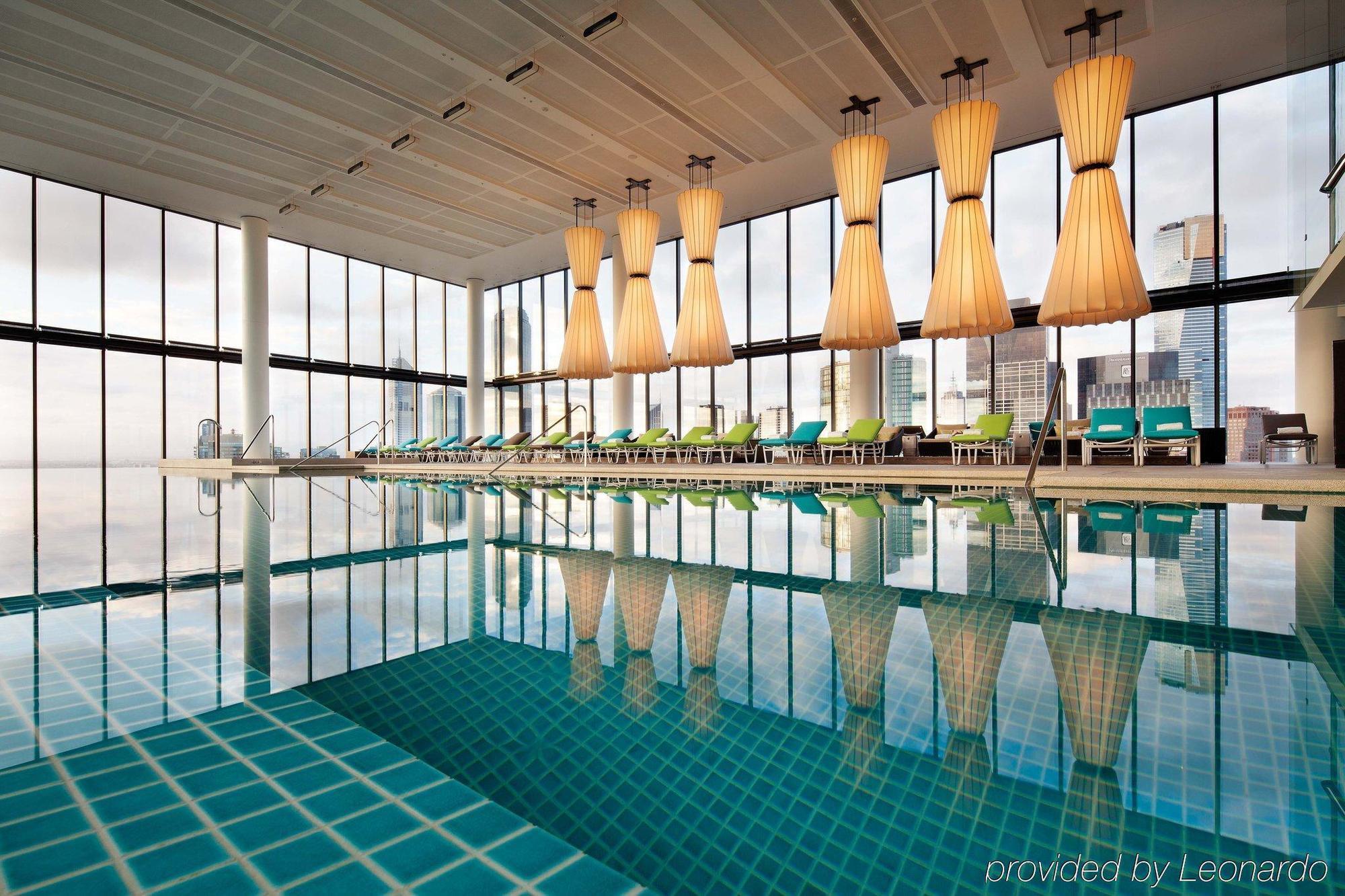 Hotel Crown Metropol Melbourne Esterno foto The photo shows a modern indoor swimming pool area. The pool features a tiled surface and reflects large, stylish light fixtures hanging from the ceiling. Surrounding the pool are several lounge chairs arranged neatly, and the walls consist of large 