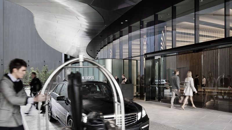 Hotel Crown Metropol Melbourne Esterno foto The image shows a modern hotel entrance with sleek architecture. In the foreground, there is a black luxury car parked near a curved entryway. A luggage cart is positioned to the side, indicating a welcoming environment for guests. People are seen wa