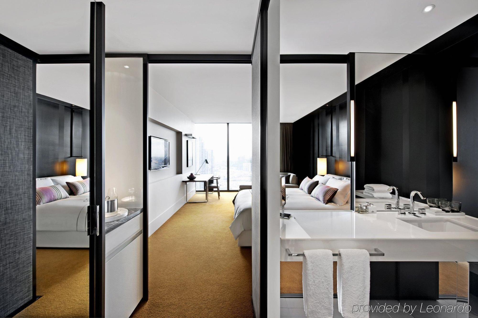 Hotel Crown Metropol Melbourne Esterno foto The photo shows a modern hotel room with a sleek design. On the left side, there is a large bed with two bedside lamps, complemented by wall-mounted artwork. Straight ahead, a large window allows natural light to illuminate the room, and a desk is po