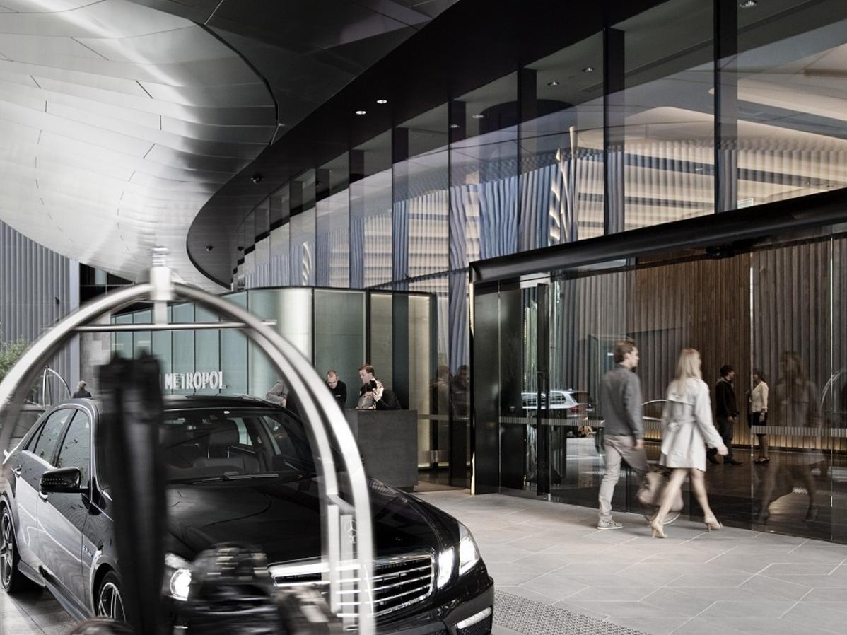 Hotel Crown Metropol Melbourne Esterno foto The photo depicts a modern hotel entrance featuring a sleek design. In the foreground, a black car is parked near a circular driveway. People are seen entering and exiting the building, which has a spacious and stylish facade characterized by large g