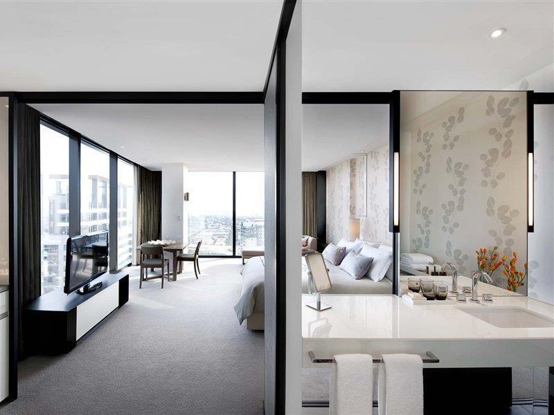 Hotel Crown Metropol Melbourne Esterno foto The image shows a modern hotel room or suite featuring a spacious layout divided by a glass or partition wall. On one side, there is a bedroom area with a large bed, and the decor appears stylish with neutral colors. The opposite side includes a livi