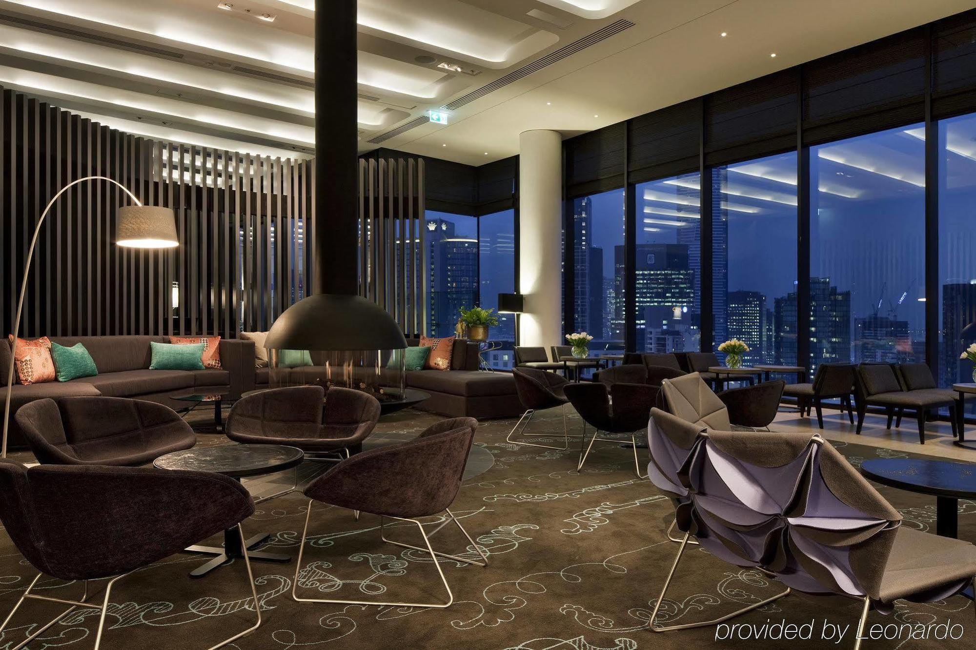 Hotel Crown Metropol Melbourne Esterno foto The image depicts a modern and stylish lounge area. The space features large windows that provide a view of a city skyline at dusk. The interior is designed with a combination of dark and light elements, including a sleek fireplace in the center. The