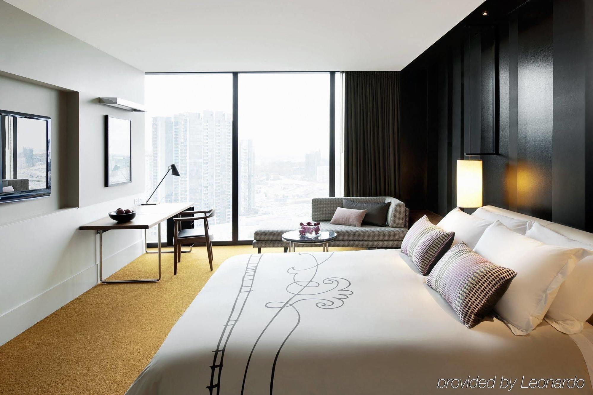 Hotel Crown Metropol Melbourne Camera foto The photo shows a modern hotel room featuring a large bed with decorative pillows, situated against a dark wall. There is a desk with a chair next to a window that offers a view of the cityscape. A small seating area includes a couch and a round tabl