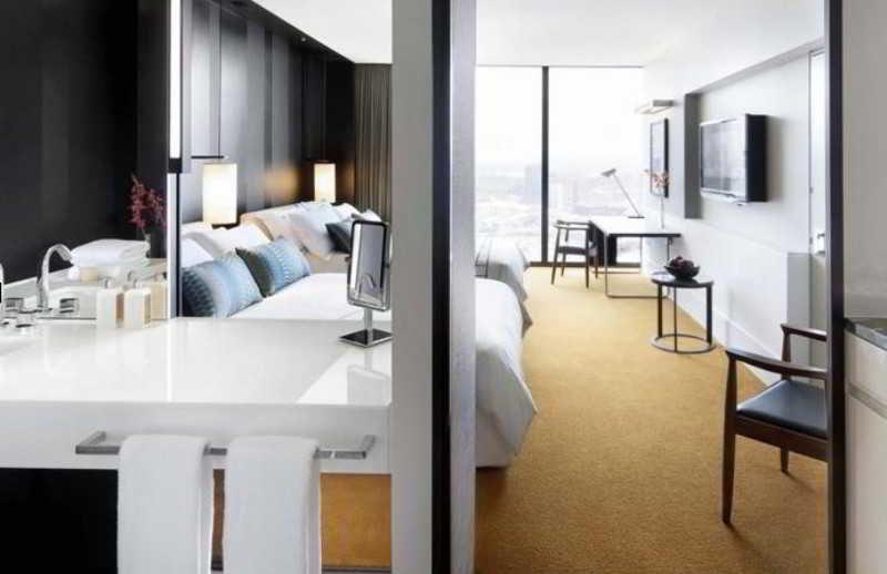 Hotel Crown Metropol Melbourne Esterno foto The photo shows a modern hotel room divided into two sections. On the left side, there are two beds with plush bedding and several decorative pillows. A sink and a small countertop area suggest a separate space for personal grooming. On the right sid