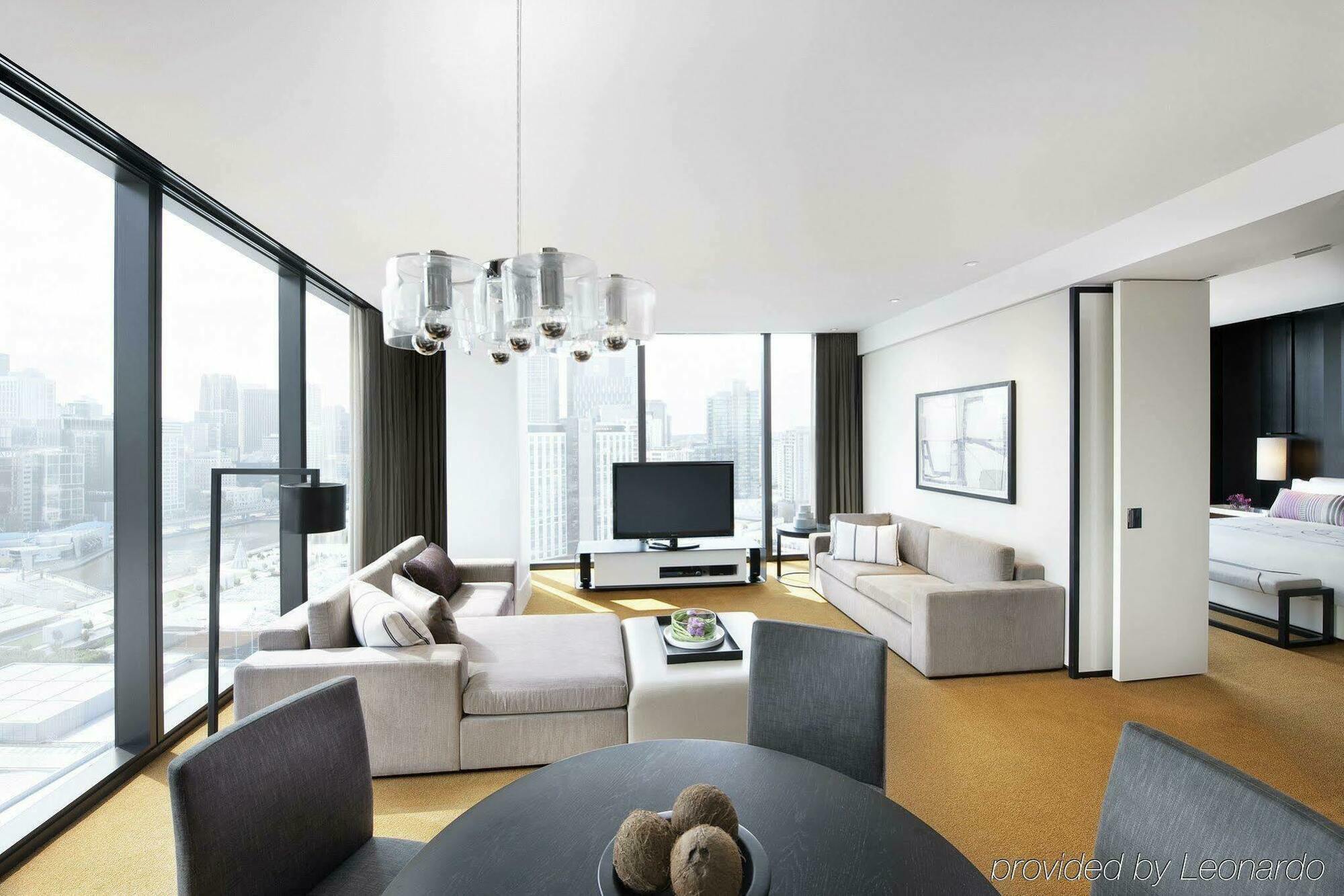 Hotel Crown Metropol Melbourne Camera foto The photo shows a modern, stylish living space. The room features large windows that provide a panoramic view of the city skyline. There is a comfortable light-colored sofa arranged in the corner, complemented by a dark floor lamp. Across from the so