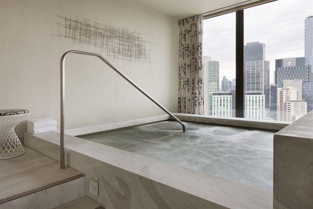 Hotel Crown Metropol Melbourne Esterno foto The photo features a modern indoor hot tub or spa located near a large window with a view of city skyscrapers. The tub is rectangular and has a metallic safety railing. The surrounding area is designed with sleek, neutral-colored materials, and there